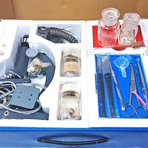 May include: A vintage microscope kit with various scientific tools and equipment. The kit includes a gray microscope, glass jars, and dissection tools, all neatly arranged in a blue box with a white interior.