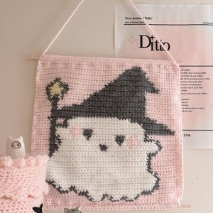 May include: A square crochet wall hanging featuring a white ghost character wearing a dark gray witch hat and holding a yellow star-topped wand. The ghost is set against a light pink background, and the piece is suspended by a matching yarn string.