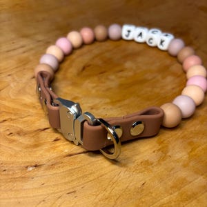 Può includere: Un collare per cani in pelle marrone con una fibbia argentata e un anello. Il collare è decorato con perline di silicone rosa e beige e ha il nome "JACK" scritto in lettere bianche.