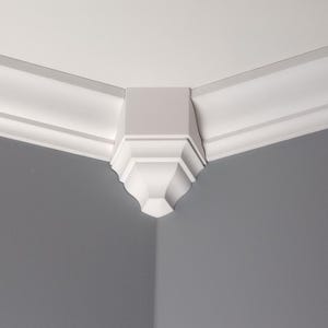 Puede incluir: Una esquina de habitación con moldura de corona blanca y una pieza de esquina decorativa. La moldura tiene un diseño en capas, y la pieza de esquina tiene un borde inferior curvo y detallado. Las paredes están pintadas de gris neutro.