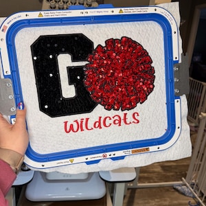 May include: A white fabric square held in a blue 11x13 inch embroidery hoop featuring a large black letter G with white sequins, a red sequined pom-pom shape, and the word Wildcats embroidered in red thread below the letter and pom-pom.