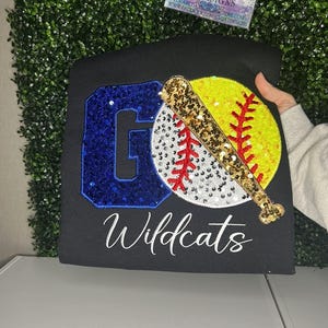May include: Black t-shirt with a sequined design featuring a blue letter "G", a yellow and white softball with red stitching, and a gold sequined baseball bat. The word "Wildcats" is written in white script.