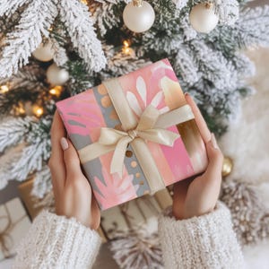 May include: A wrapped gift with pink, grey, and gold floral patterned paper and a cream-coloured ribbon. The gift is held in front of a Christmas tree with white baubles and lights.