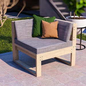 May include: A modern outdoor corner chair with a light wood frame and gray cushions. Two throw pillows in green and tan add a pop of color. The chair is on a brick patio, with greenery in the background.