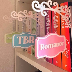 Può includere: Cartello decorativo bianco per scaffali con tre etichette appese: "TBR", "Romance" e un'altra con testo illeggibile. I cartelli sono in colori pastello e sono appesi a una staffa bianca decorativa. I libri sono visibili dietro i cartelli.