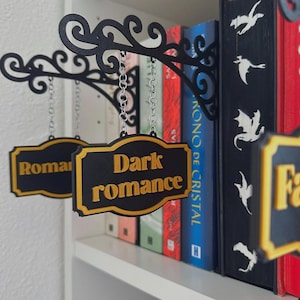 Op de afbeelding: Zwarte en gouden decoratieve borden hangen voor een boekenplank. De borden lezen "Dark romance" en "Romance" in gouden letters. De borden zijn opgehangen aan kettingen van sierlijke zwarte beugels.