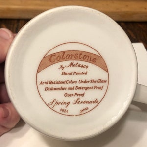 May include: A white ceramic mug with a brown label that reads "Colorstone by Metasco Hand Painted Acid Resistant Colors Under The Glaze Dishwasher and Detergent Proof Oven Proof Spring Serenade 4024 JAPAN".