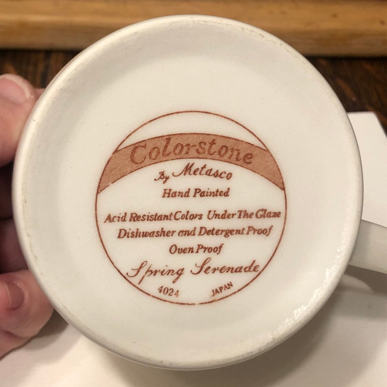May include: A white ceramic mug with a brown label that reads "Colorstone by Metasco Hand Painted Acid Resistant Colors Under The Glaze Dishwasher and Detergent Proof Oven Proof Spring Serenade 4024 JAPAN".