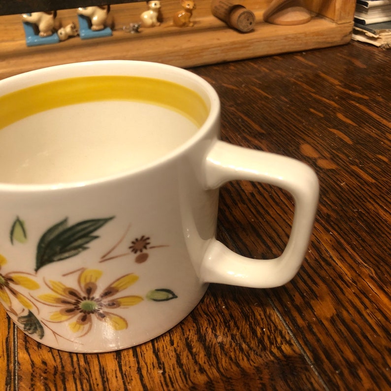 May include: A white ceramic mug with a yellow rim and a floral design featuring brown and yellow flowers and green leaves. The mug has a white handle.