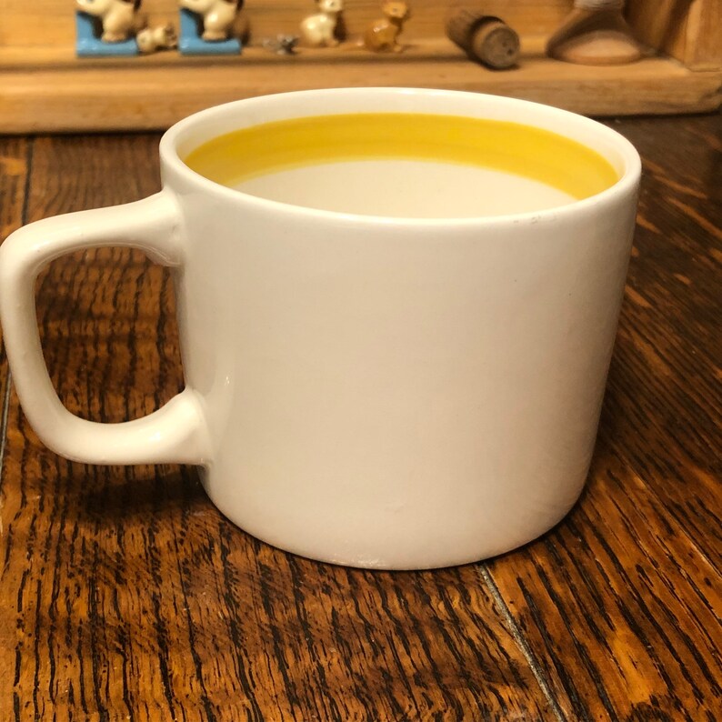 May include: A white ceramic mug with a yellow rim, filled with a light-colored beverage. The mug is sitting on a wooden surface.