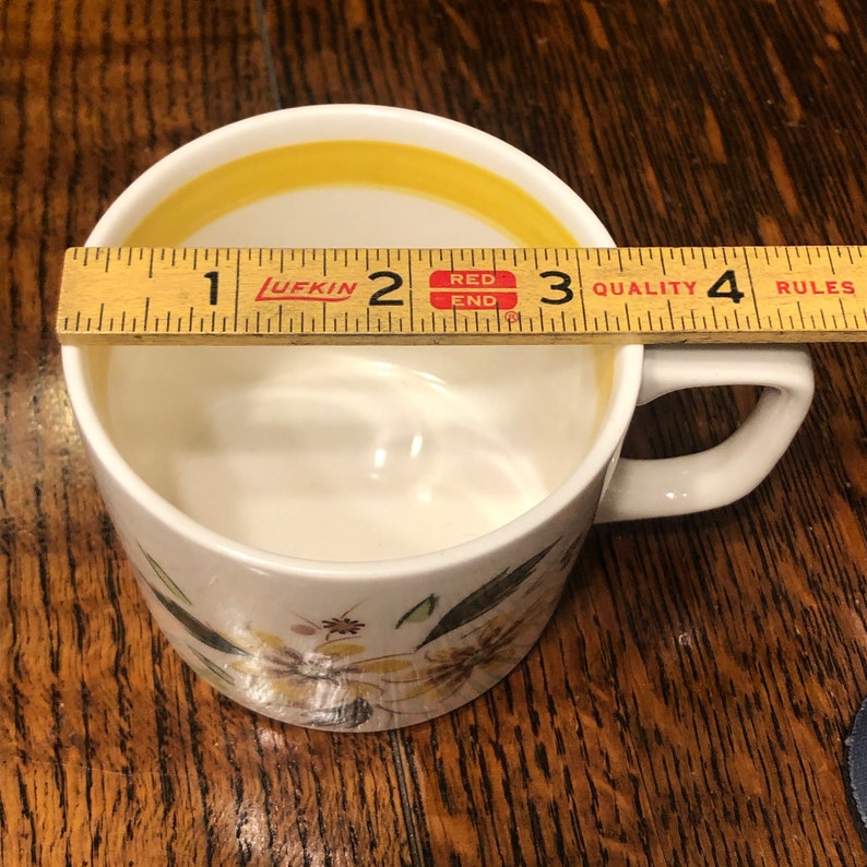 May include: A white ceramic mug with a yellow rim and floral pattern. The mug is 2.5 inches tall and has a handle. The mug is sitting on a wooden surface.
