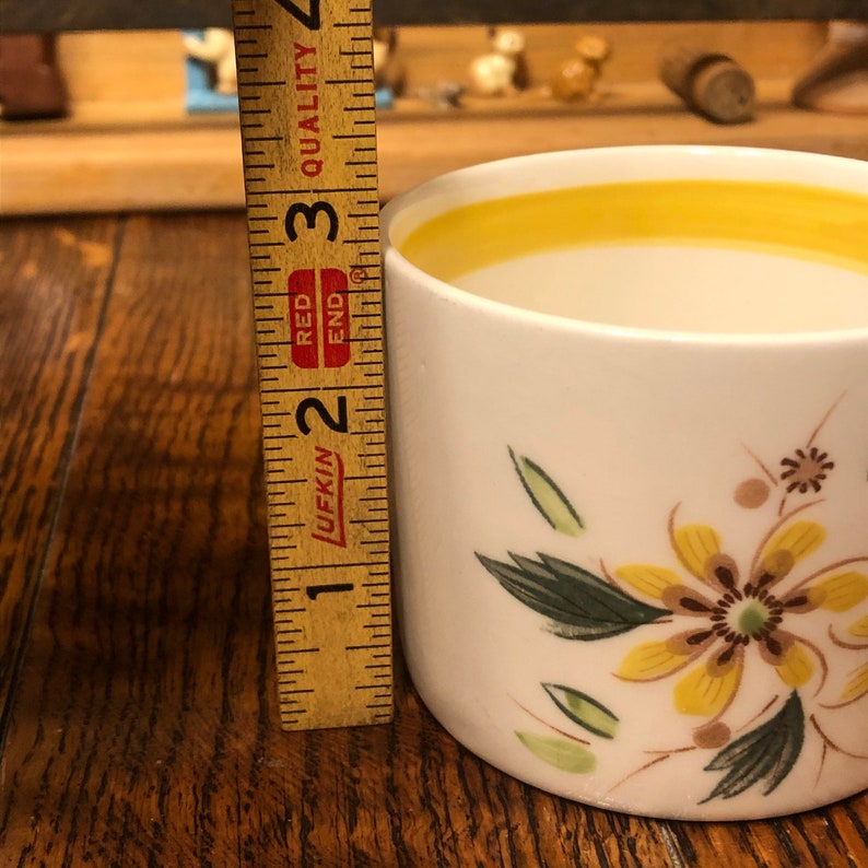 May include: A white ceramic mug with a yellow rim and floral design. The mug is 2 3/4 inches tall and features a yellow flower with brown center and green leaves.