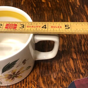 May include: White ceramic mug with a floral design and a yellow rim. The mug is 3 inches tall and has a handle. The mug is sitting on a wooden surface.