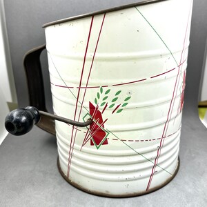 May include: White metal flour sifter with a red and green geometric design. The sifter has a crank handle and a black knob.
