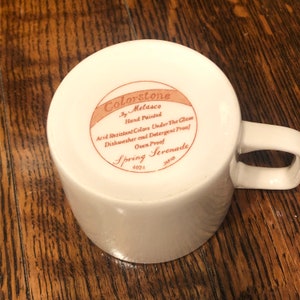 May include: White ceramic mug with a brown label that reads "Colorstone by Metasco Hand Painted Acid Resistant Colors Under The Glaze Dishwasher and Detergent Proof Oven Proof Spring Serenade 4024 JAPAN".
