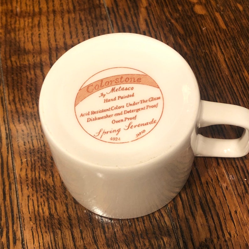 May include: White ceramic mug with a brown label that reads "Colorstone by Metasco Hand Painted Acid Resistant Colors Under The Glaze Dishwasher and Detergent Proof Oven Proof Spring Serenade 4024 JAPAN".