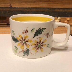 May include: A white ceramic mug with a yellow rim and a floral design featuring yellow and brown flowers with green leaves.