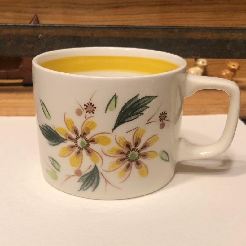 May include: A white ceramic mug with a yellow rim and a floral design featuring yellow and brown flowers with green leaves.