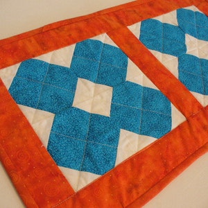 May include: A quilted table runner with a white background and two blue diamond patterns. The runner has a bright orange border with a swirl pattern.