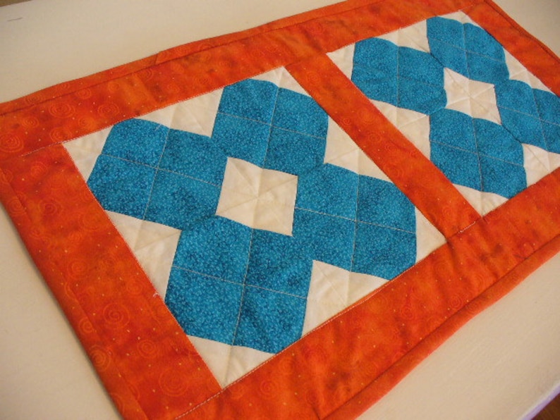 Puede incluir: Un camino de mesa acolchado con fondo blanco y dos patrones de diamantes azules. El camino de mesa tiene un borde naranja brillante con un patr&oacute;n de espiral.