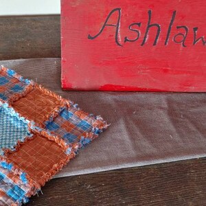 May include: A small, square, quilted cushion with a patchwork design in shades of orange and blue. The cushion is made of fabric with a frayed edge. The cushion is sitting on a white, sheer fabric.