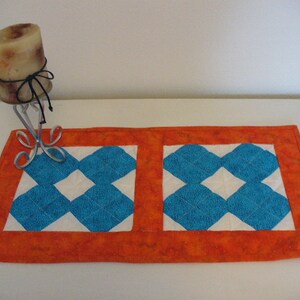 Puede incluir: Un camino de mesa acolchado naranja y blanco con un patr&oacute;n de diamantes azules. El camino de mesa est&aacute; hecho de tela y tiene un borde decorativo.