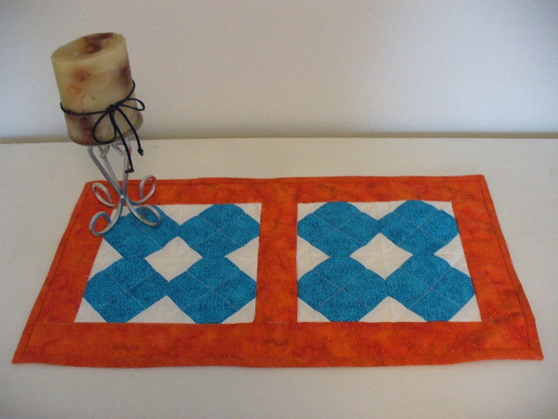 Puede incluir: Un camino de mesa acolchado naranja y blanco con un patr&oacute;n de diamantes azules. El camino de mesa est&aacute; hecho de tela y tiene un borde decorativo.