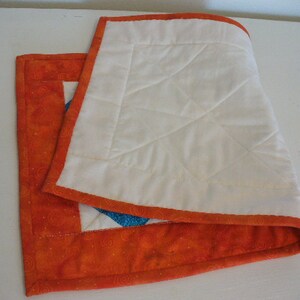 Puede incluir: Un camino de mesa acolchado blanco con un borde naranja y un patr&oacute;n de espiral. El camino de mesa est&aacute; doblado por la mitad y se encuentra sobre una superficie blanca.