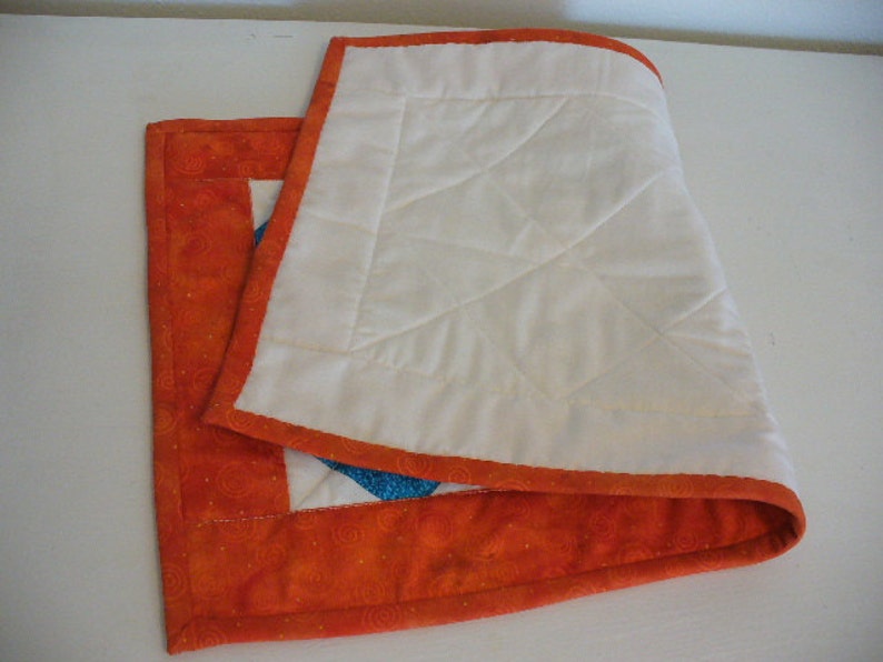 Puede incluir: Un camino de mesa acolchado blanco con un borde naranja y un patr&oacute;n de espiral. El camino de mesa est&aacute; doblado por la mitad y se encuentra sobre una superficie blanca.