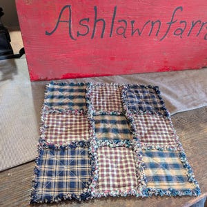 May include: A square rag quilt potholder featuring a nine-patch grid pattern. The fabric squares alternate between navy blue, tan, and brown plaid designs. Each square is stitched with an X pattern and finished with frayed, raw-edge seams.