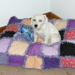 May include: A white dog with a fluffy coat is lying on a patchwork dog bed. The bed is made of various fabrics in shades of purple, pink, and brown. The bed has a ruffled edge.
