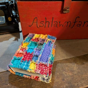May include: A colorful, handmade book cover with a patchwork design. The cover features squares of various fabrics in shades of red, blue, yellow, and purple. The text "Ashlawn farms" is visible on a red wooden box in the background. A vintage Singer sewing machine is also in the background.