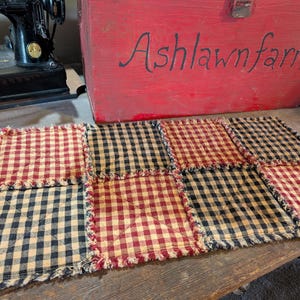 May include: A rectangular fabric mat featuring a patchwork design of alternating red and black gingham squares. The mat has frayed, raw edges between each square and is quilted with a simple cross-stitch pattern on each individual fabric block.