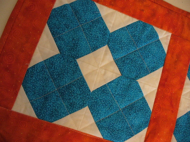 Puede incluir: Un cuadrado de tela acolchado con un patr&oacute;n geom&eacute;trico de cuadrados azul turquesa y crema. La tela est&aacute; bordeada con una tela naranja s&oacute;lida con un patr&oacute;n de espiral.