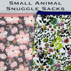 May include: Two small animal snuggle sacks. One sack features a gray background with large pink floral patterns and a pink fleece trim. The other sack has a white background with a floral and flamingo pattern and a blue fleece trim.