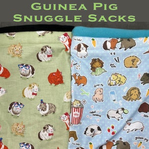 May include: Two guinea pig snuggle sacks. One is green with a black trim and features a white guinea pig pattern. The other is blue with a turquoise trim and features a variety of guinea pig patterns, including a guinea pig eating popcorn, a guinea pig wearing a dress, and a guinea pig with a bottle of water.