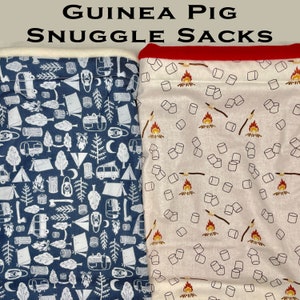 May include: Two guinea pig snuggle sacks. One is navy blue with a white camping theme print. The other is off-white with a campfire and marshmallow print. The top of one sack is red, and the other is white. The text "GUINEA PIG SNUGGLE SACKS" is at the top.