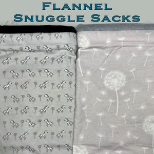 Pode incluir: Dois sacos de flanela para aconchegar. Um apresenta um padrão de zebras e palmeiras em um fundo cinza claro, o outro tem um design de dente-de-leão com sementes brancas em um fundo cinza claro. A parte superior do saco de zebra é preta e o saco de dente-de-leão é cinza.