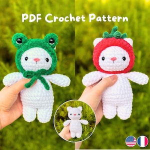 Puede incluir: Dos muñecos de ganchillo blancos, uno con un gorro de rana verde y otro con un gorro de fresa rojo. Los muñecos tienen ojos negros y narices rosas. La imagen incluye el texto "PDF Crochet Pattern".
