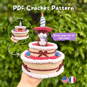 May include: A crocheted birthday cake featuring an otter emerging from the top. The cake is two-tiered, with red, white, and beige sections, adorned with blue berries and brown bows. A blue and white candle is on top. Text reads "PDF Crochet Pattern".