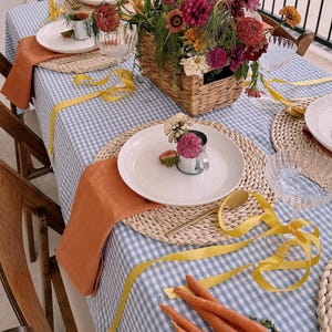 Op de afbeelding: Een gedekte tafel met een blauw-wit geblokt tafelkleed, met borden, geweven placemats, oranje servetten en een mand met kleurrijke bloemen. Gele linten en wortelen voegen decoratieve accenten toe.
