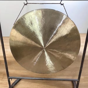 Puede incluir: Un gran gong circular de bronce suspendido dentro de un marco de metal oscuro. La superficie del gong refleja la luz, mostrando un patrón radial desde el centro. El marco tiene un diseño geométrico simple, adecuado para la meditación o la música.