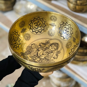 May include: A golden singing bowl with intricate etched designs. The bowl features the Om symbol and an image of a deity. The bowl is held by a person wearing a black long-sleeve shirt.