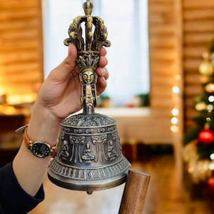 Może przedstawiać: Ozdobny, brązowy dzwon z misternymi rzeźbami postaci i wzorów. Dzwon ma dekoracyjny uchwyt z motywem przypominającym twarz. Dzwon trzymany jest w dłoni, co podkreśla jego kunszt i design.