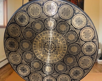May include: A large, round gong with an intricate, symmetrical design in gold and black. The gong is suspended from a black metal bar and features a central geometric pattern surrounded by circular motifs. The overall aesthetic is ornate and spiritual.