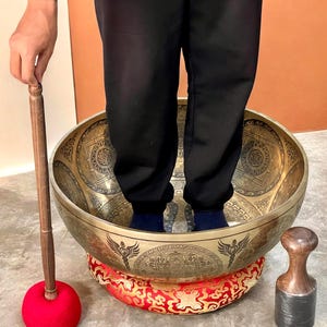 Può includere: Una grande e ornata ciotola tibetana in ottone con intricati disegni incisi. La ciotola poggia su un cuscino a fantasia rosso e oro. Una persona è in piedi all'interno della ciotola. Sono presenti anche una mazza di legno con testa in feltro rosso e un percussore in legno.