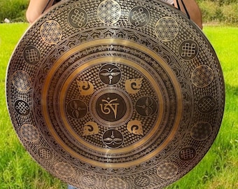 May include: A large, circular bronze gong with intricate etched patterns. The design includes geometric shapes, symbols, and a central Om symbol. The gong is held by black straps, and the background is a green grassy area.