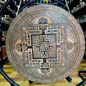 May include: A large, round, brass gong with a detailed mandala design in gold and black. The gong is hanging from a stand.