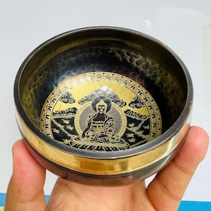 Puede incluir: Un cuenco cantor negro con un borde dorado y un diseño de Buda dorado en el interior. El cuenco está decorado con nubes y pájaros.