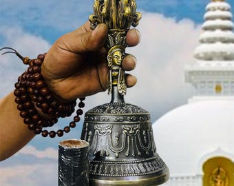 May include: A hand holds a silver and gold bell with intricate carvings and a small figure on the top. The bell is attached to a black wooden handle. The background is a white stupa with a golden finial.